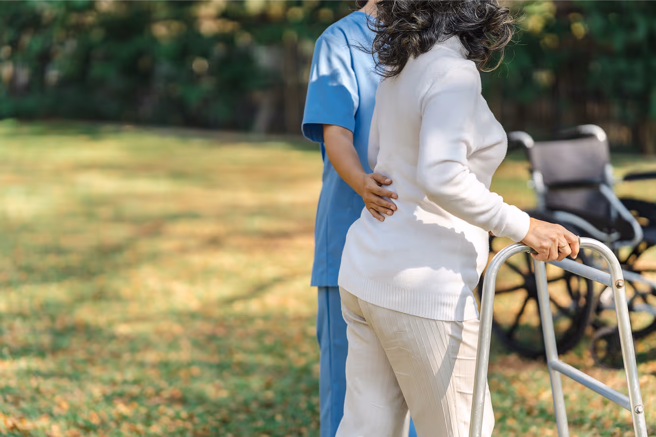 Caregiver in blue scrubs supporting an elderly person using a walker outdoors with a wheelchair in the background.