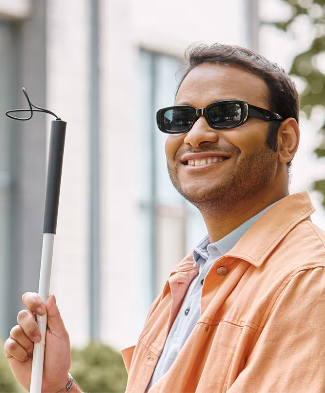 Smiling man wearing sunglasses and an orange jacket holding a white cane outdoors.