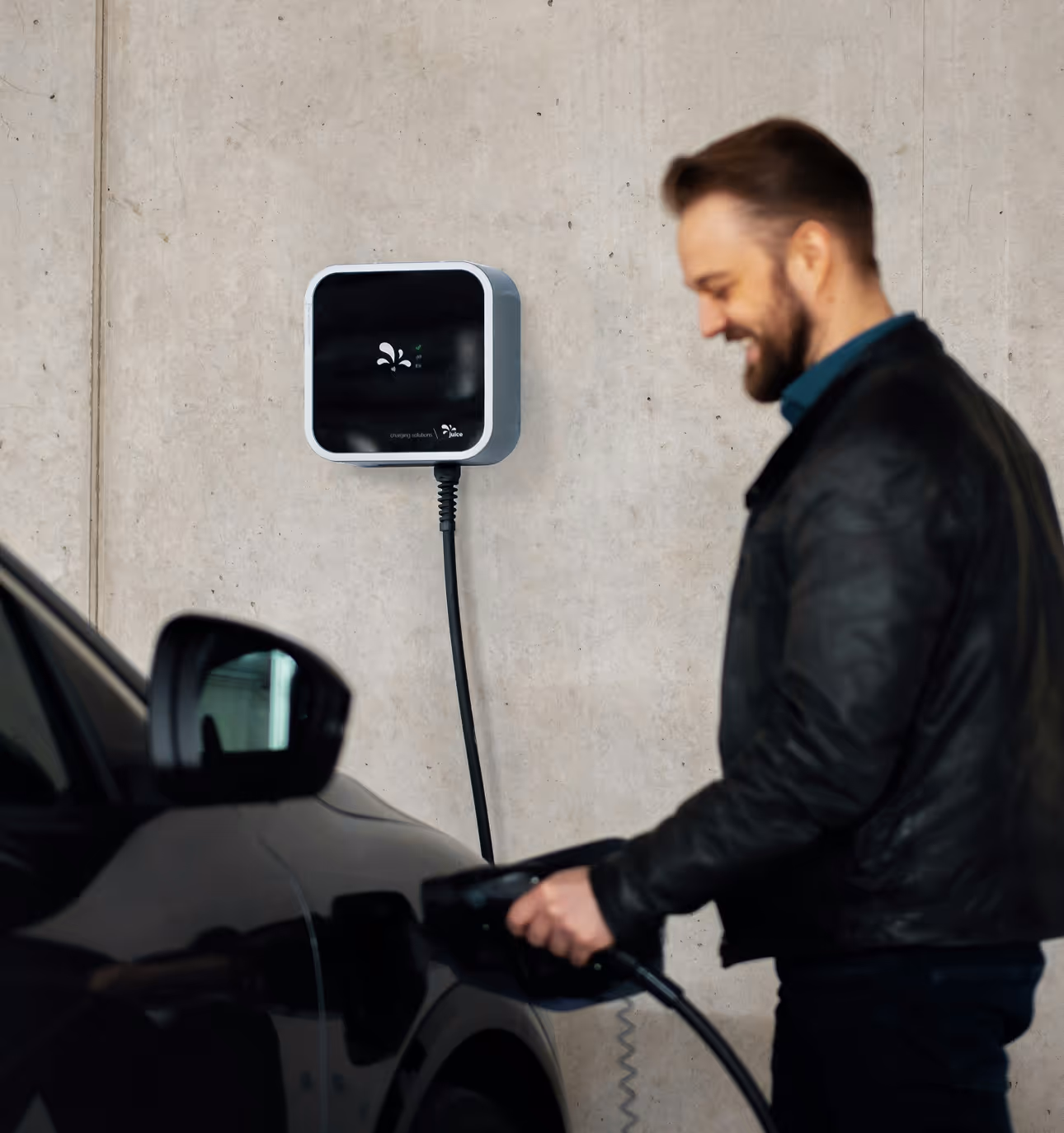 Man in black leather jacket plugging in an electric car charger to a black vehicle in front of a concrete wall.