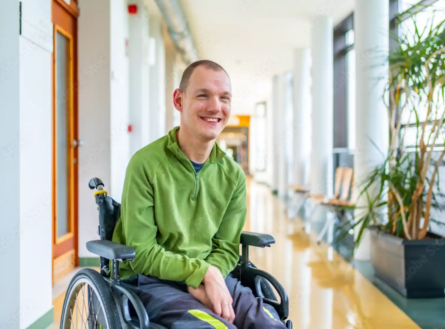 Un homme souriant en fauteuil roulant dans un couloir lumineux avec des plantes et des chaises en arrière-plan.