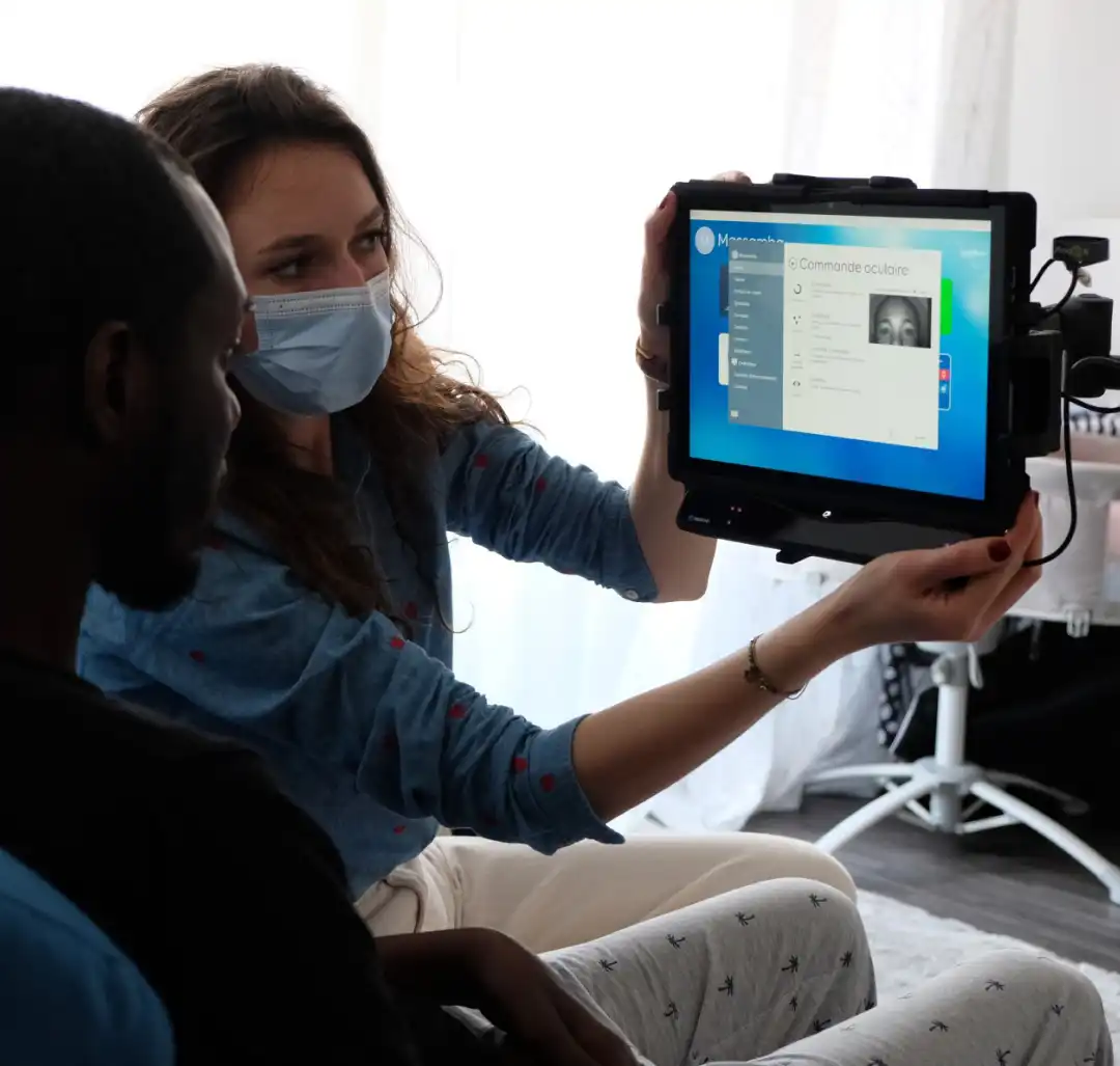 Une femme portant un masque médical montre à un homme un écran de tablette affichant un logiciel de commande oculaire.