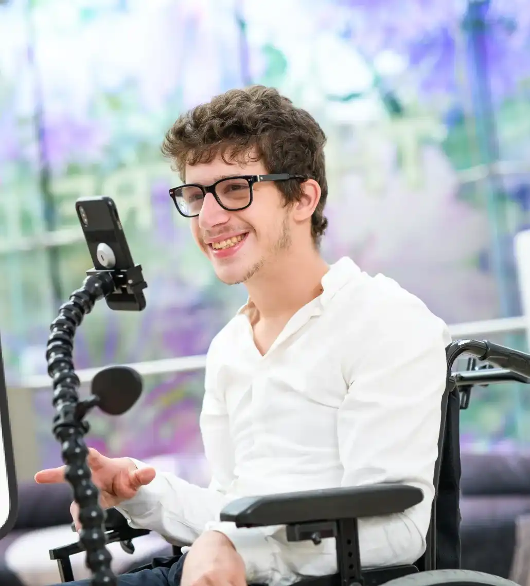 Jeune homme souriant en fauteuil roulant utilisant un smartphone fixé à un support flexible.