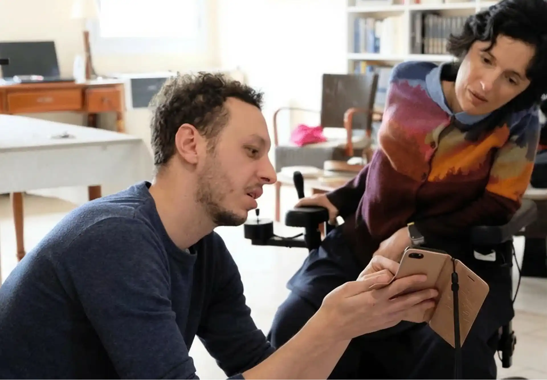 Un homme montre un téléphone portable à une femme en fauteuil roulant dans un salon avec des meubles et une bibliothèque.