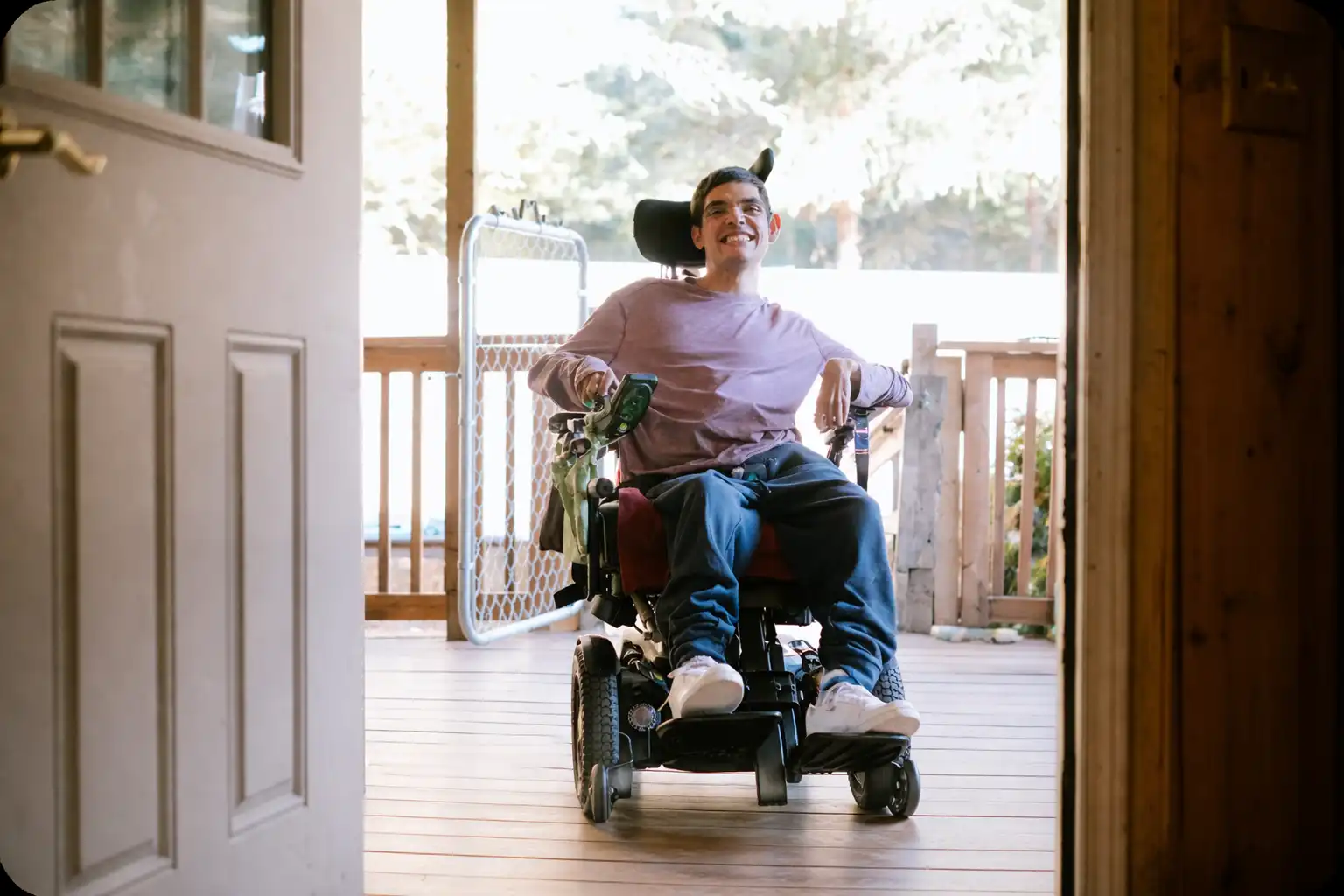 Jeune homme souriant en fauteuil roulant électrique sur une terrasse en bois avec une porte ouverte.