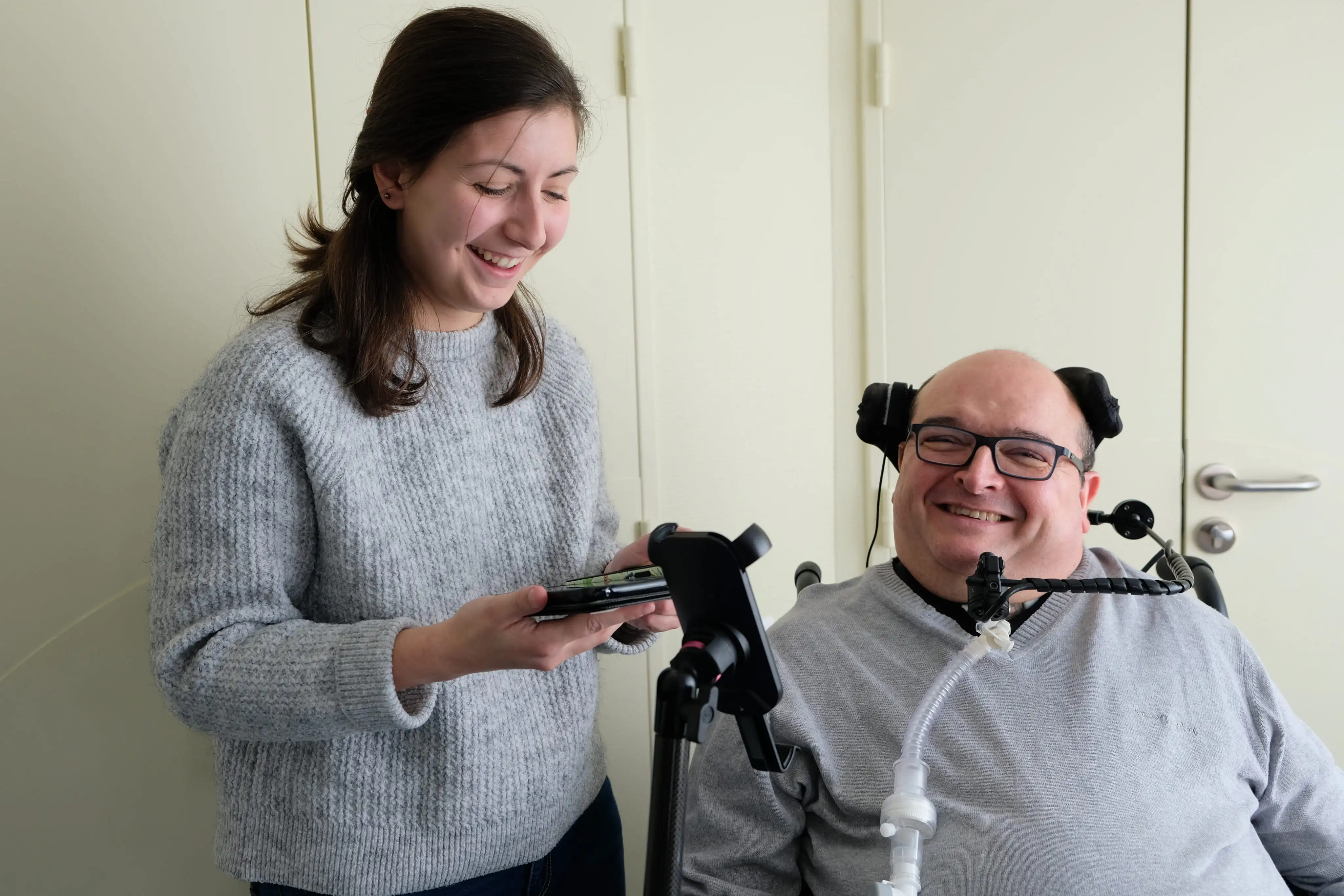 Un homme en fauteuil roulant souriant avec un appareil respiratoire, accompagné d'une femme souriante tenant un téléphone.