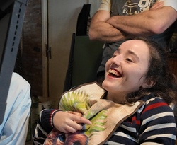 Jeune femme souriante en fauteuil roulant, portant un bavoir coloré et un pull rayé, avec une personne debout en arrière-plan.