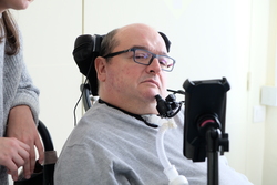 Homme en fauteuil roulant équipé d'un dispositif de communication assistée, accompagné d'une personne à côté.