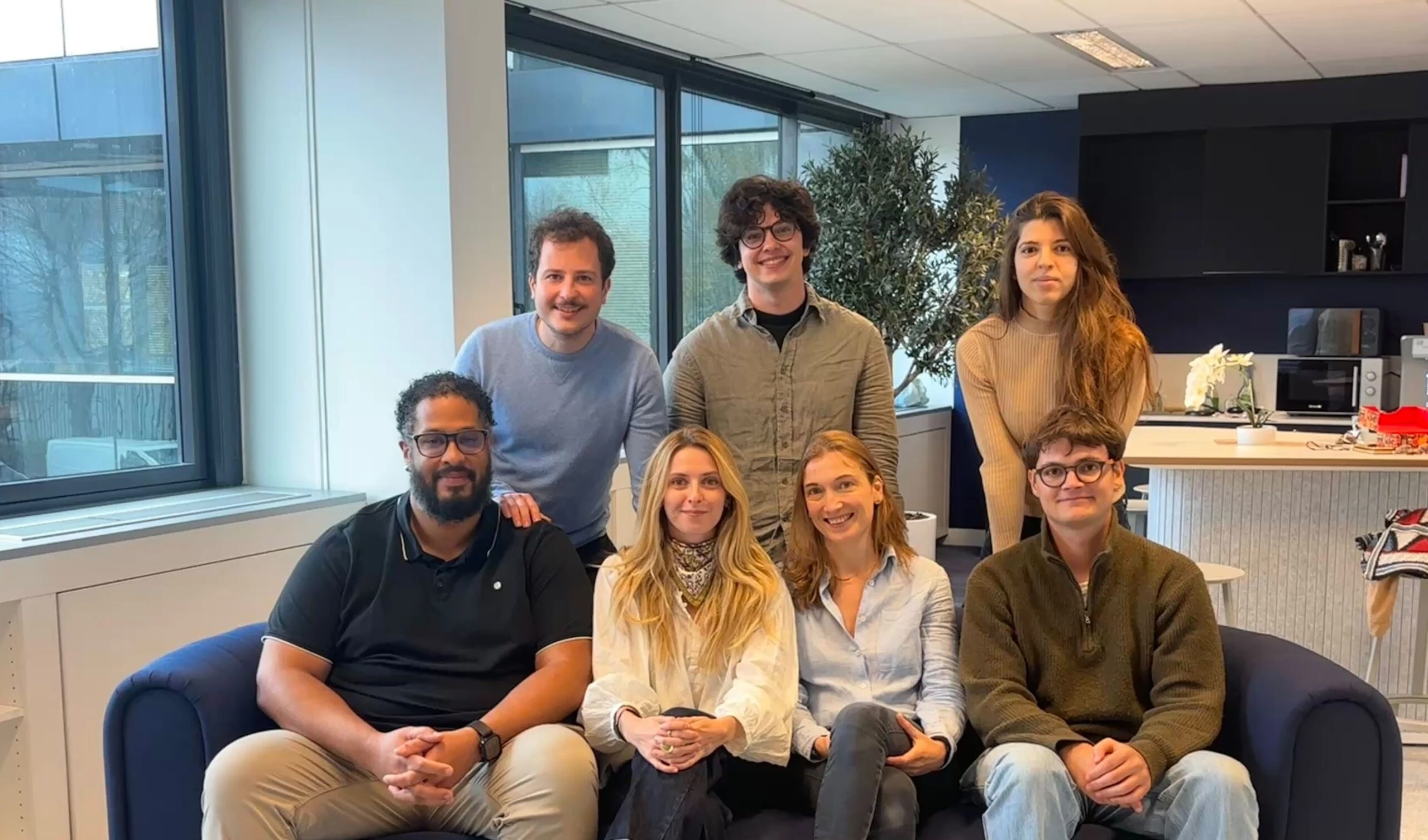Groupe de sept collègues souriants posant dans un bureau moderne avec canapé bleu et grande fenêtre.