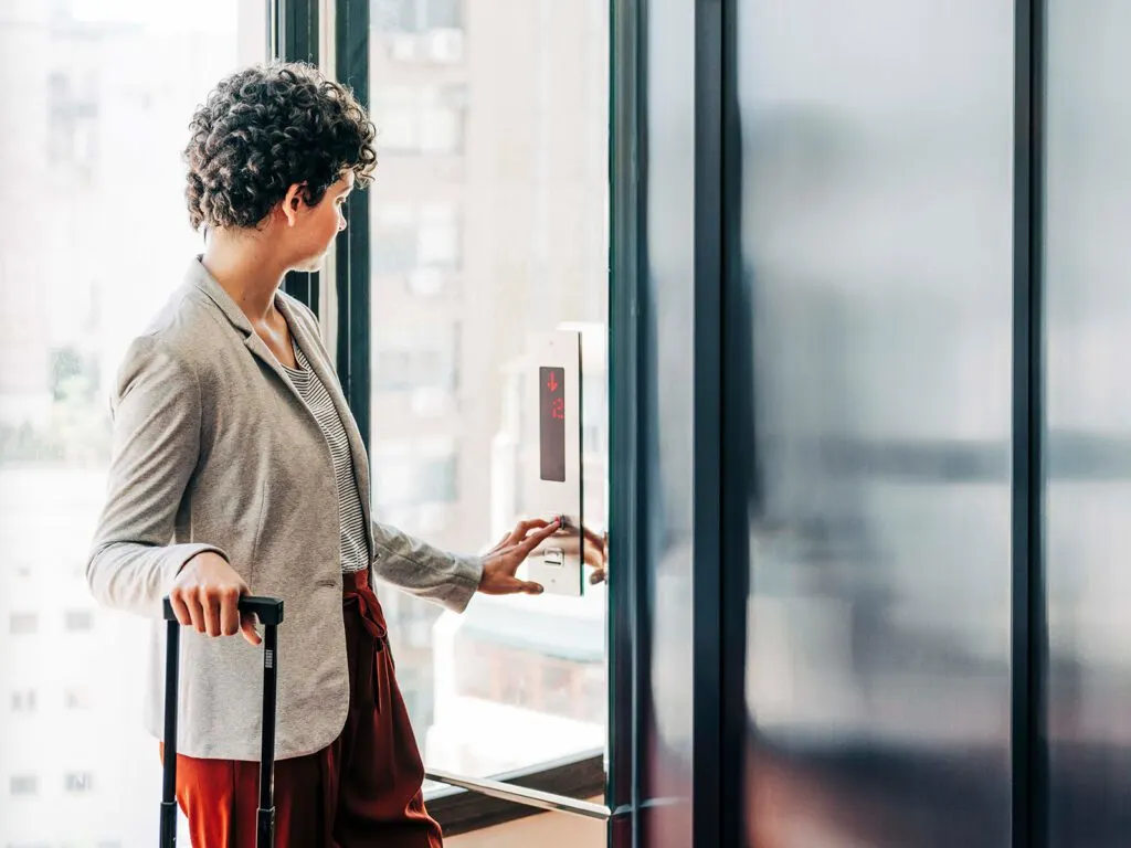 A professional woman with a suitcase using a secure keypad to access an elevator in a modern building. This image illustrates streamlined access control for residents or guests, emphasizing how RemoteLock enhances convenience and security in multifamily properties or vacation rentals.