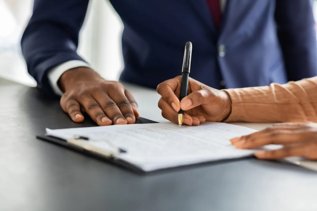 This image depicts a close-up of two people interacting over a document on a table. One individual is holding a pen and appears to be signing the document, while the other person places a hand near the document, possibly guiding or supervising. Both individuals are dressed in professional attire, suggesting a formal or business setting.
