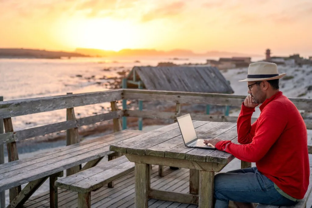 A man in a red sweater and hat works on his laptop at a rustic seaside table during sunset. This illustrates the flexibility of managing properties remotely with RemoteLock, enabling efficient access control and property operations from any location.