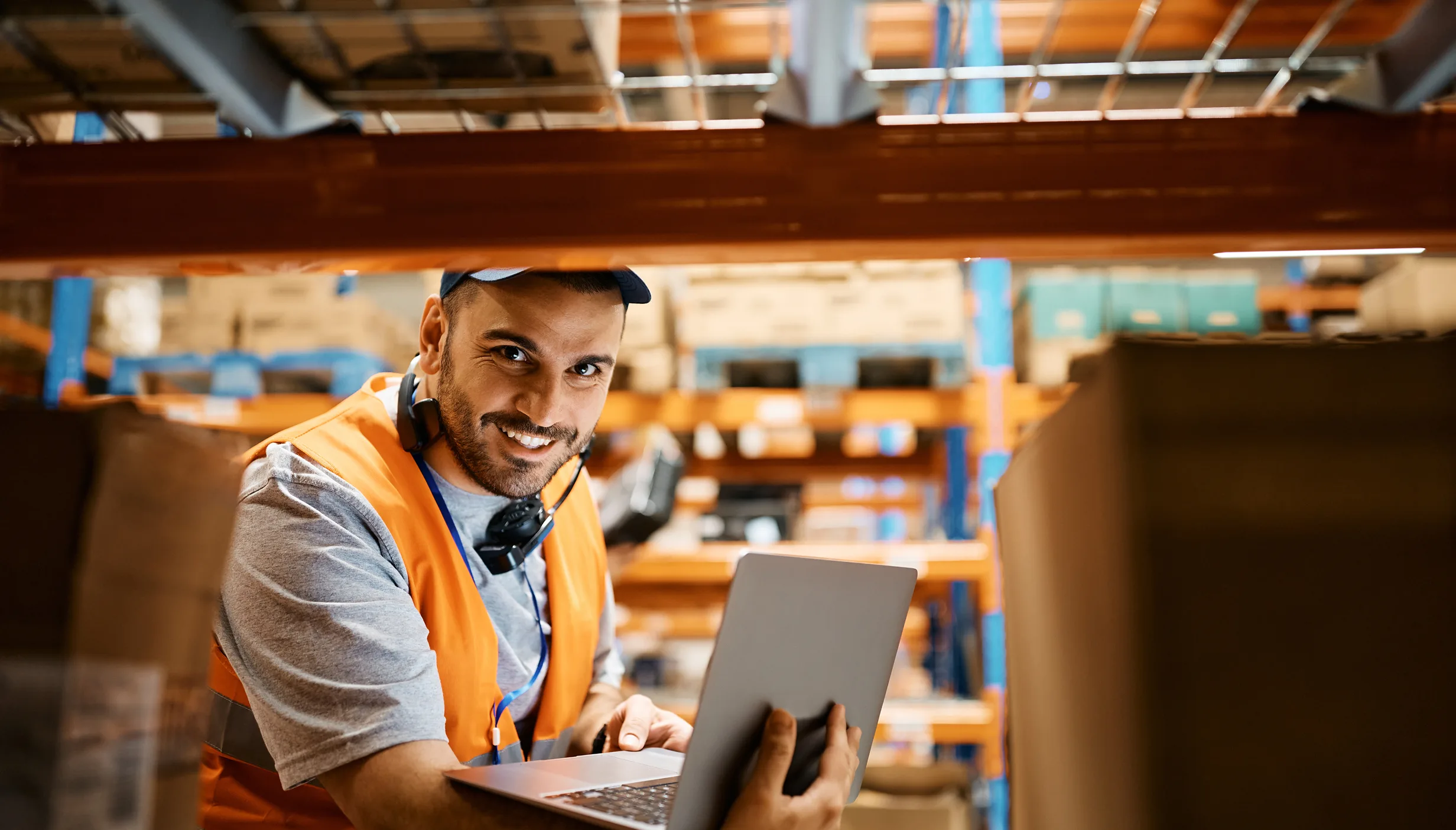 Warehouse worker wearing an orange safety vest using a laptop and smiling.