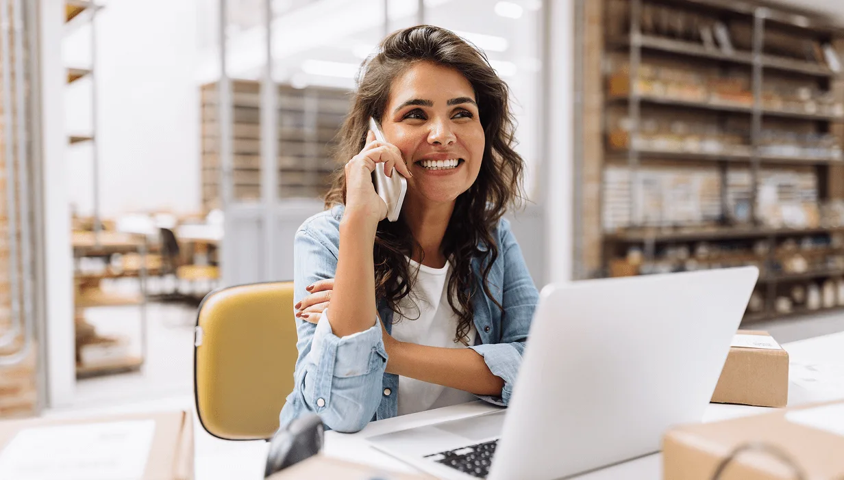 Smiling woman talking on a smartphone while sitting at a desk with a laptop in a modern office.
