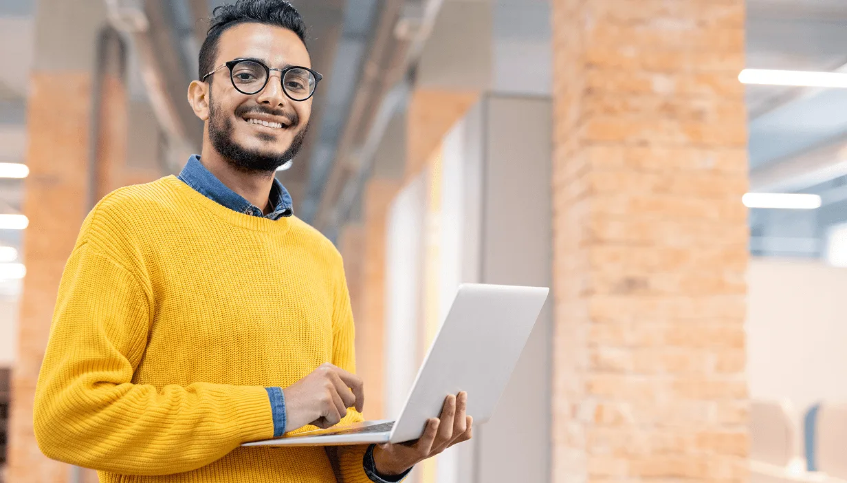 Smiling man wearing glasses and a yellow sweater holding an open laptop in a modern office.
