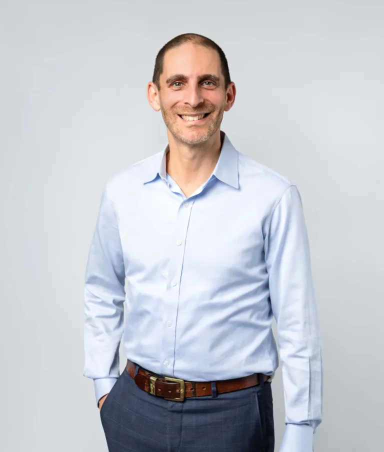 Smiling man with short hair and beard wearing a light blue button-up shirt and dark trousers against a plain light background.