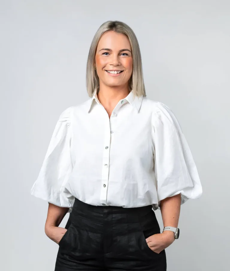 Smiling woman with blonde hair wearing a white blouse with puff sleeves and black pants, hands in pockets, standing against a plain light gray background.