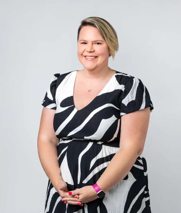 Smiling woman with short blonde hair wearing a black and white patterned dress and a pink smartwatch standing against a plain light background.