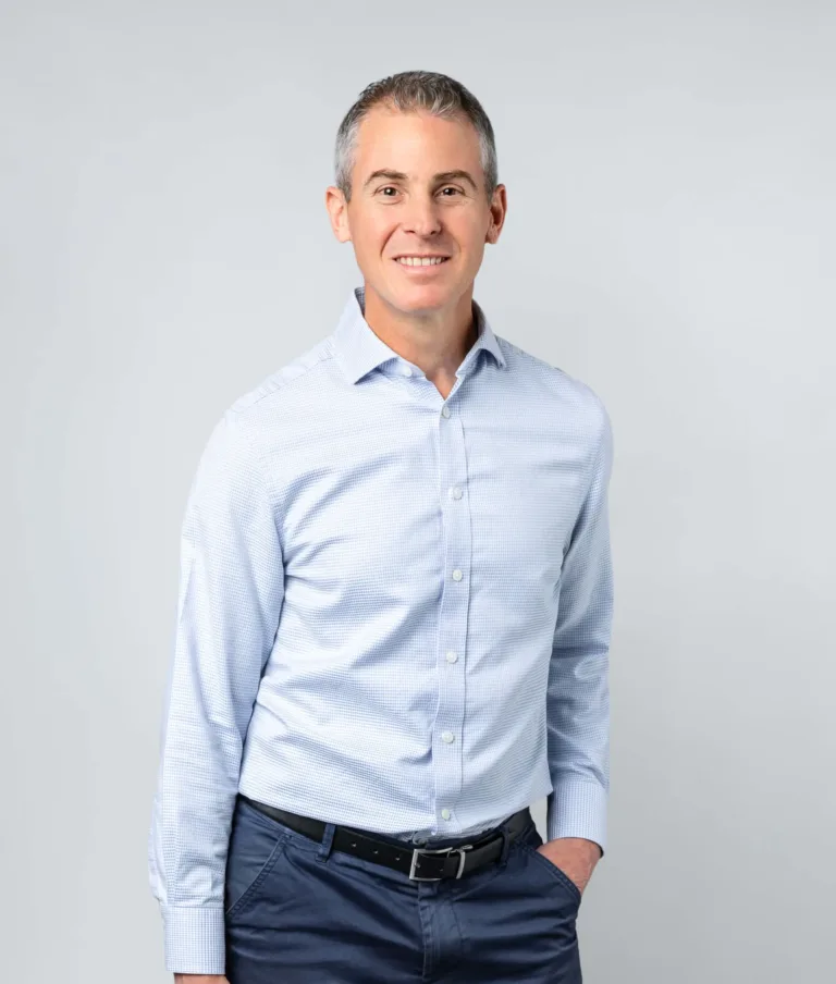 Smiling middle-aged man with short gray hair wearing a light blue dress shirt and dark pants with a black belt, standing against a light gray background.