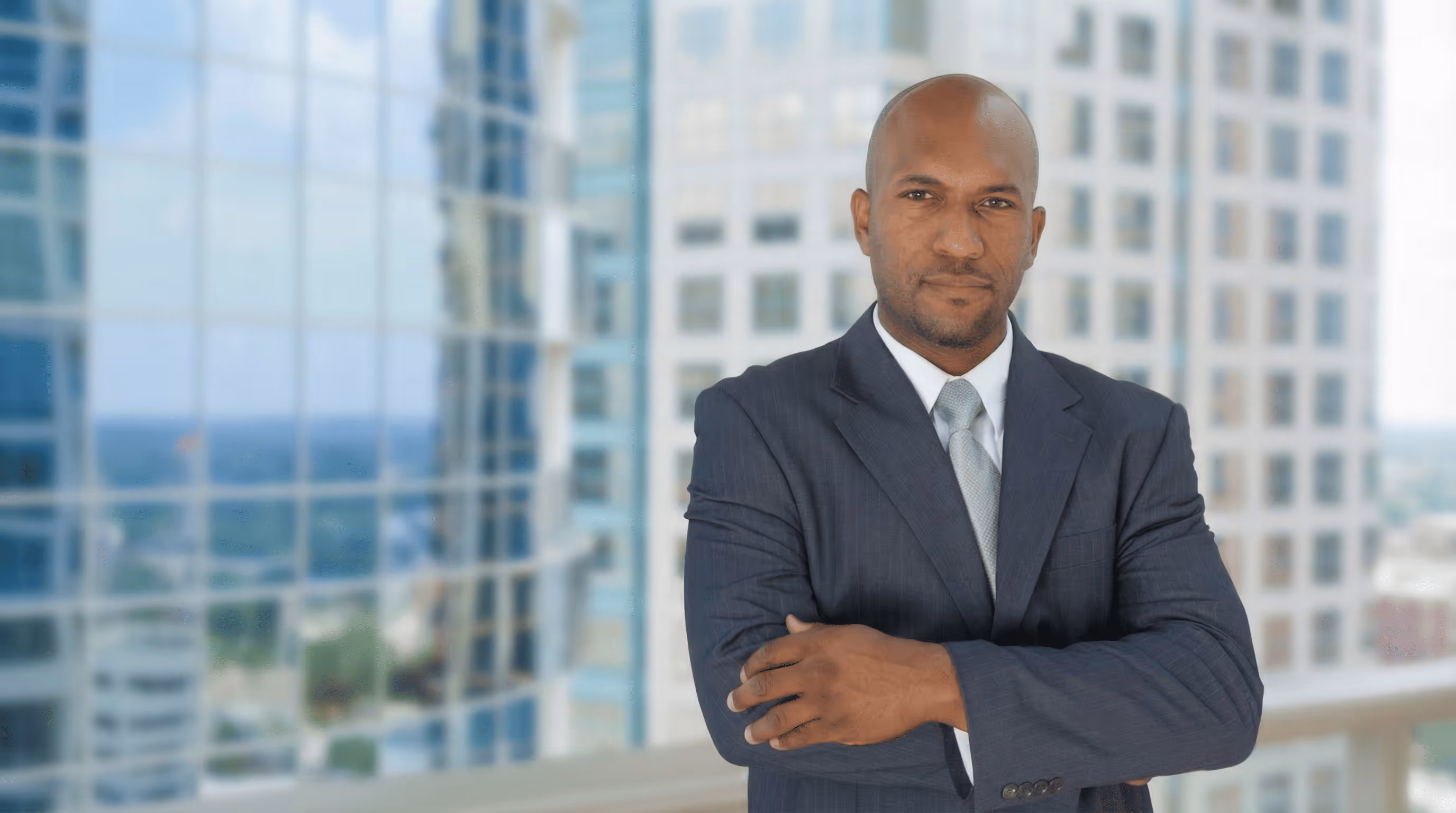 Confident businessman in a dark suit with arms crossed standing in front of a cityscape with glass buildings.