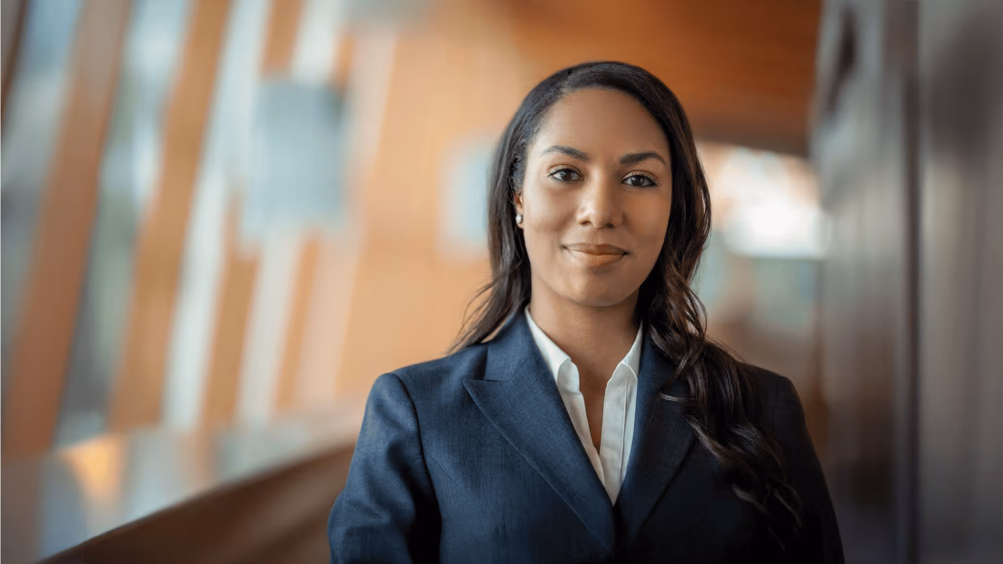 Confident woman with long dark hair wearing a navy blazer and white shirt standing indoors with blurred background.