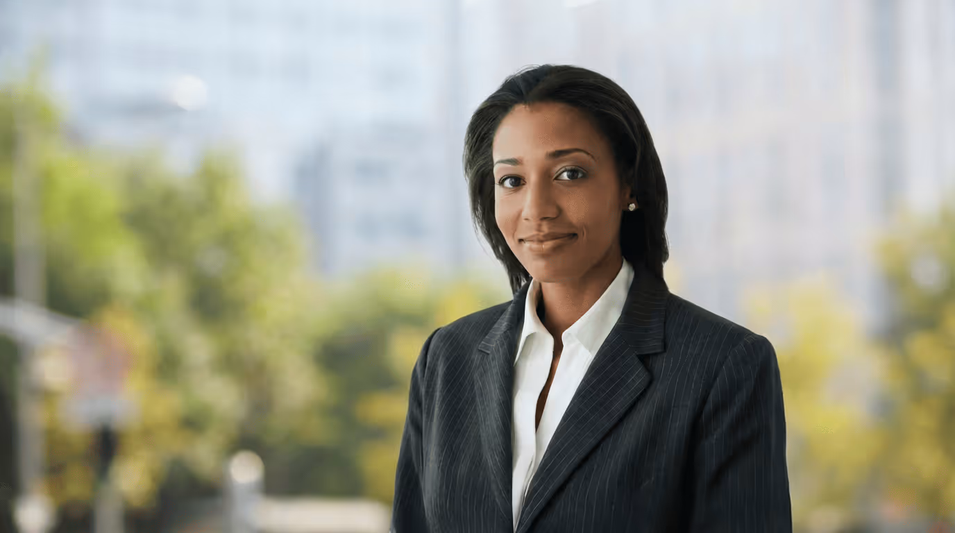 Confident businesswoman wearing a dark pinstripe suit and white blouse standing outdoors with blurred greenery and buildings in the background.