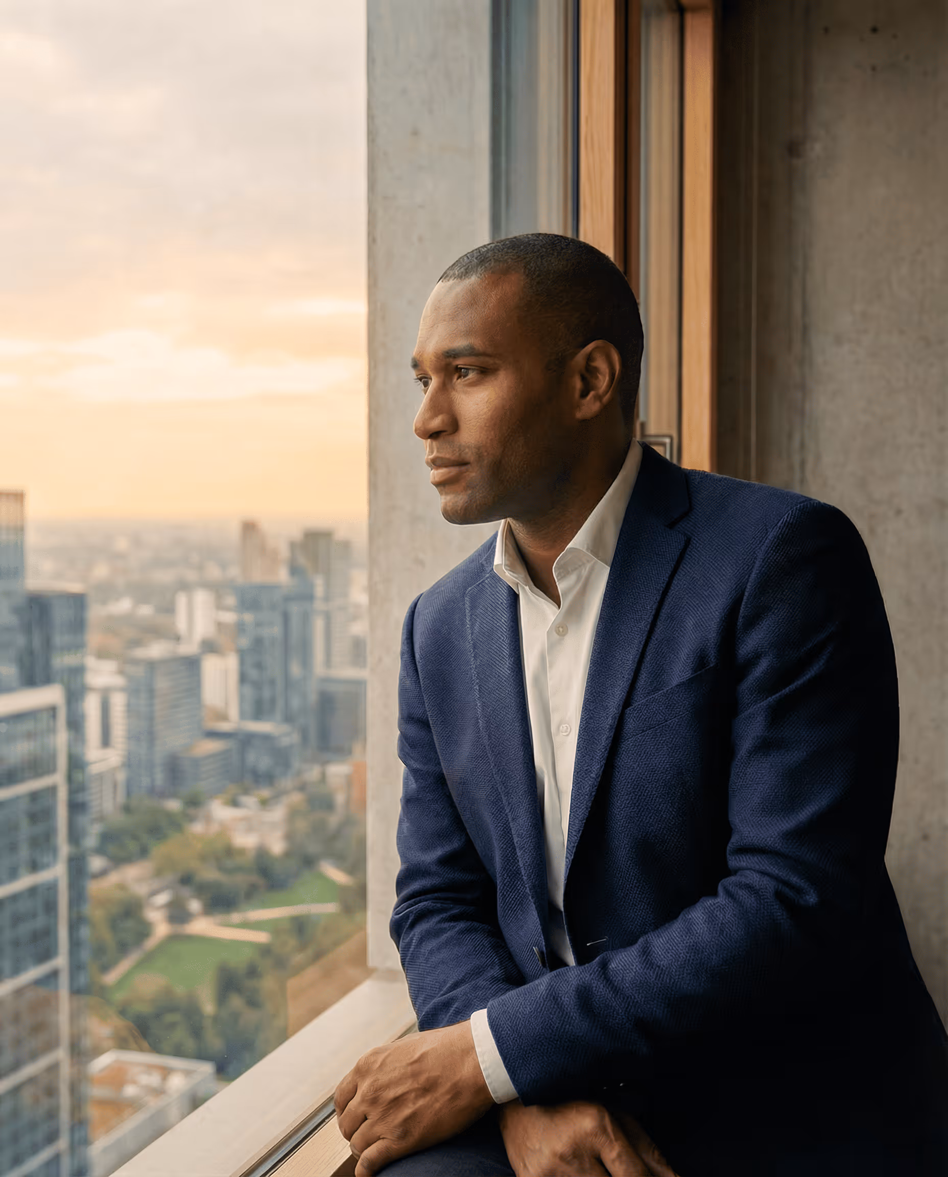Man in a navy blue suit and white shirt looking thoughtfully out a large window overlooking a cityscape at sunset.