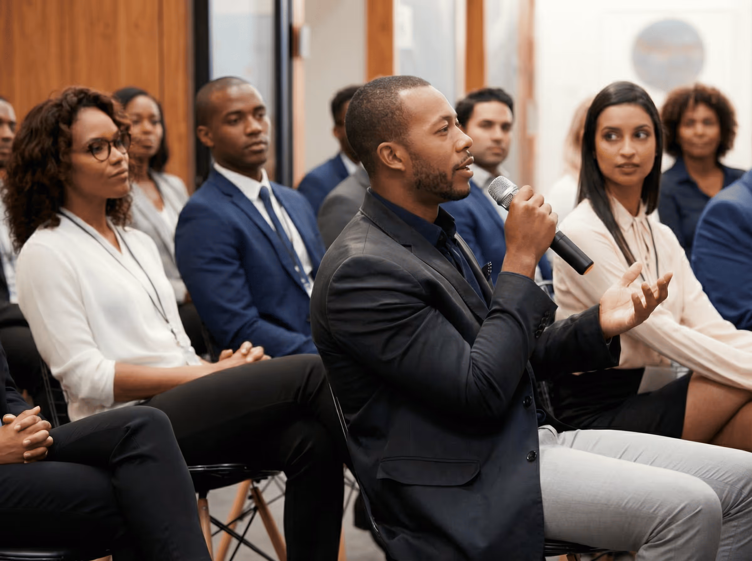 Man in a black blazer speaking into a microphone during a business meeting with attentive attendees.