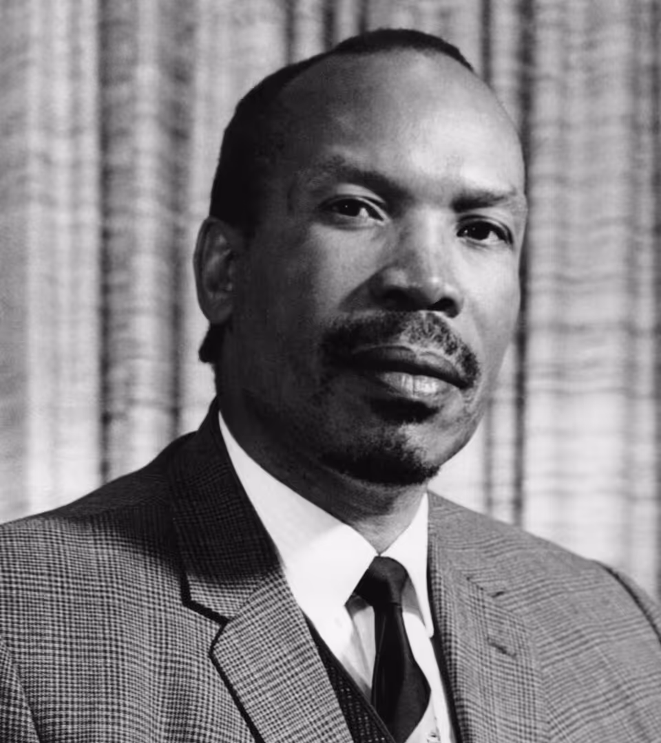 Black and white portrait of a man in a suit and tie with a calm expression against a textured curtain background.