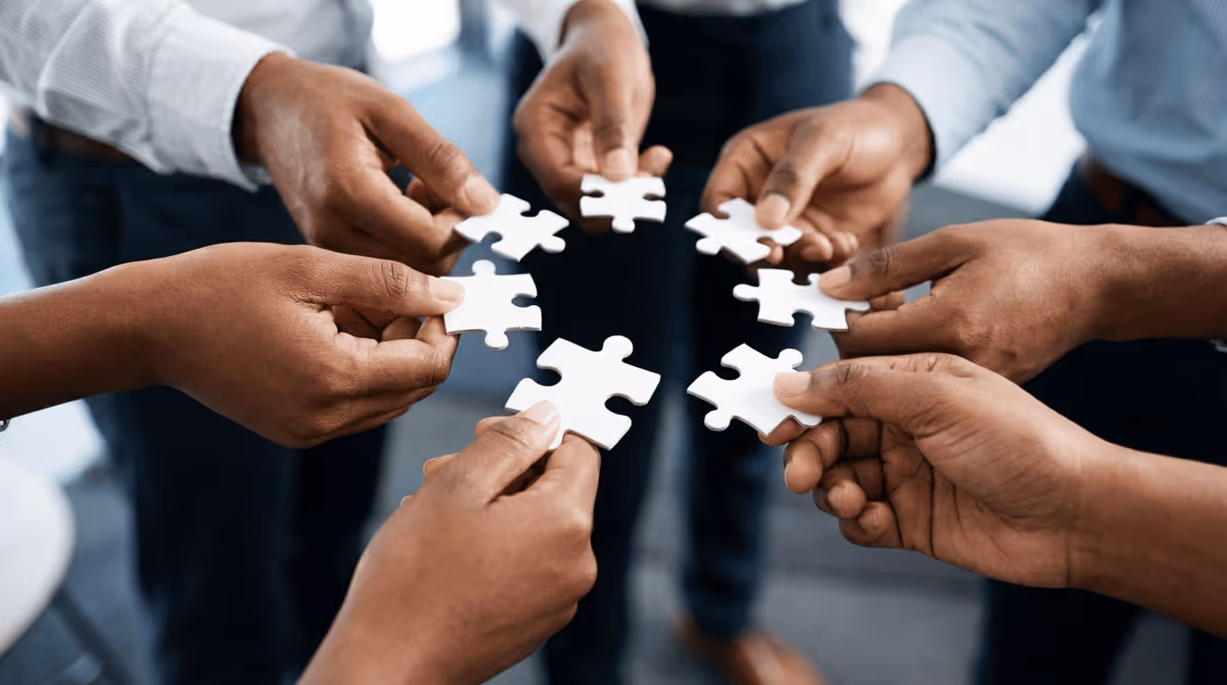 Seven people holding white puzzle pieces together in a circle, symbolizing teamwork and collaboration.