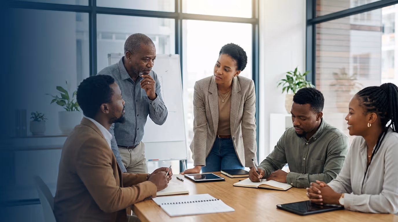 Five diverse professionals engaged in a collaborative meeting around a table in a bright office.