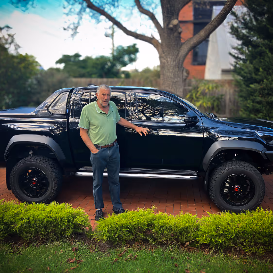 Older man in a light green polo shirt standing beside a large black pickup truck on a brick driveway.