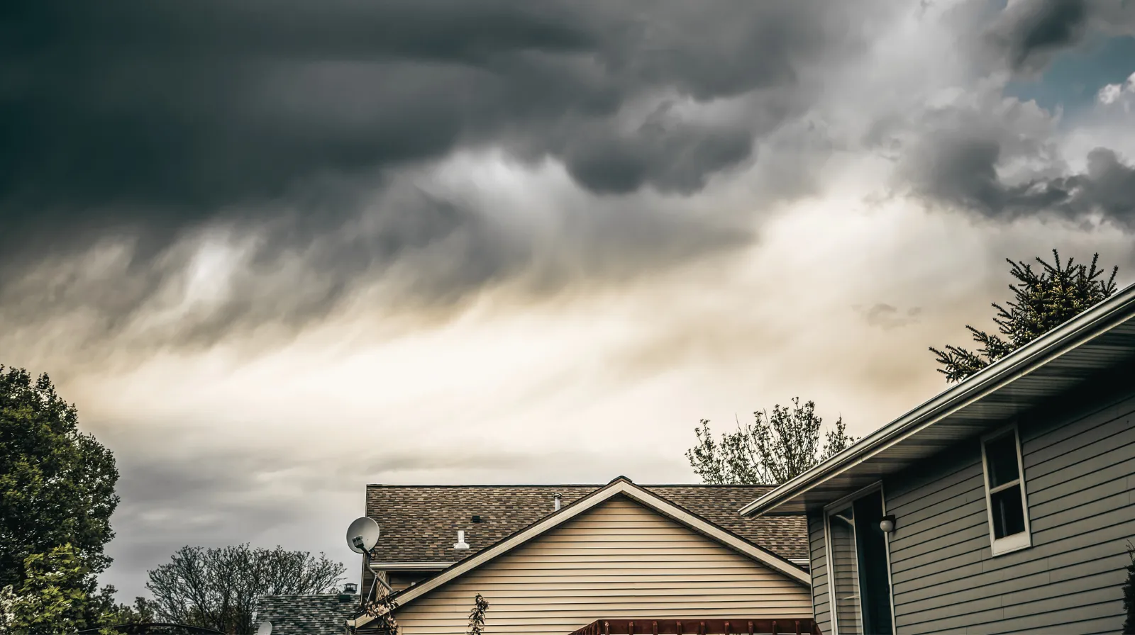 Dark storm clouds gather above suburban house rooftops and trees.