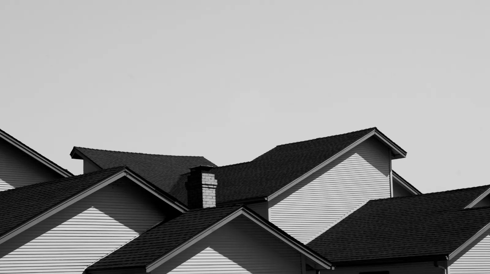 Black and white photo of several house roofs with asphalt shingles and a brick chimney under a clear sky.