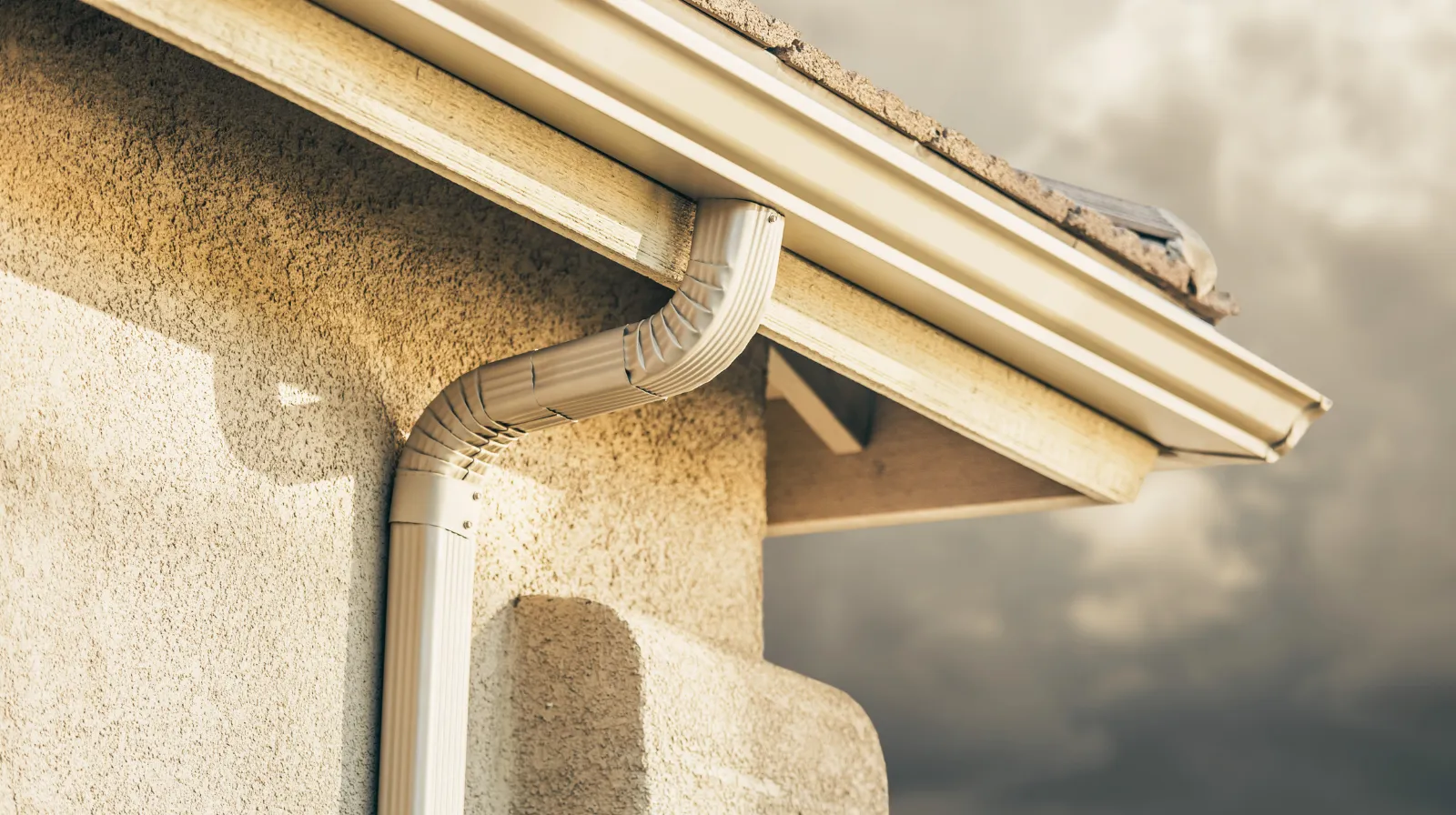 Beige house gutter system with downspout attached to stucco wall under a shingled roof.