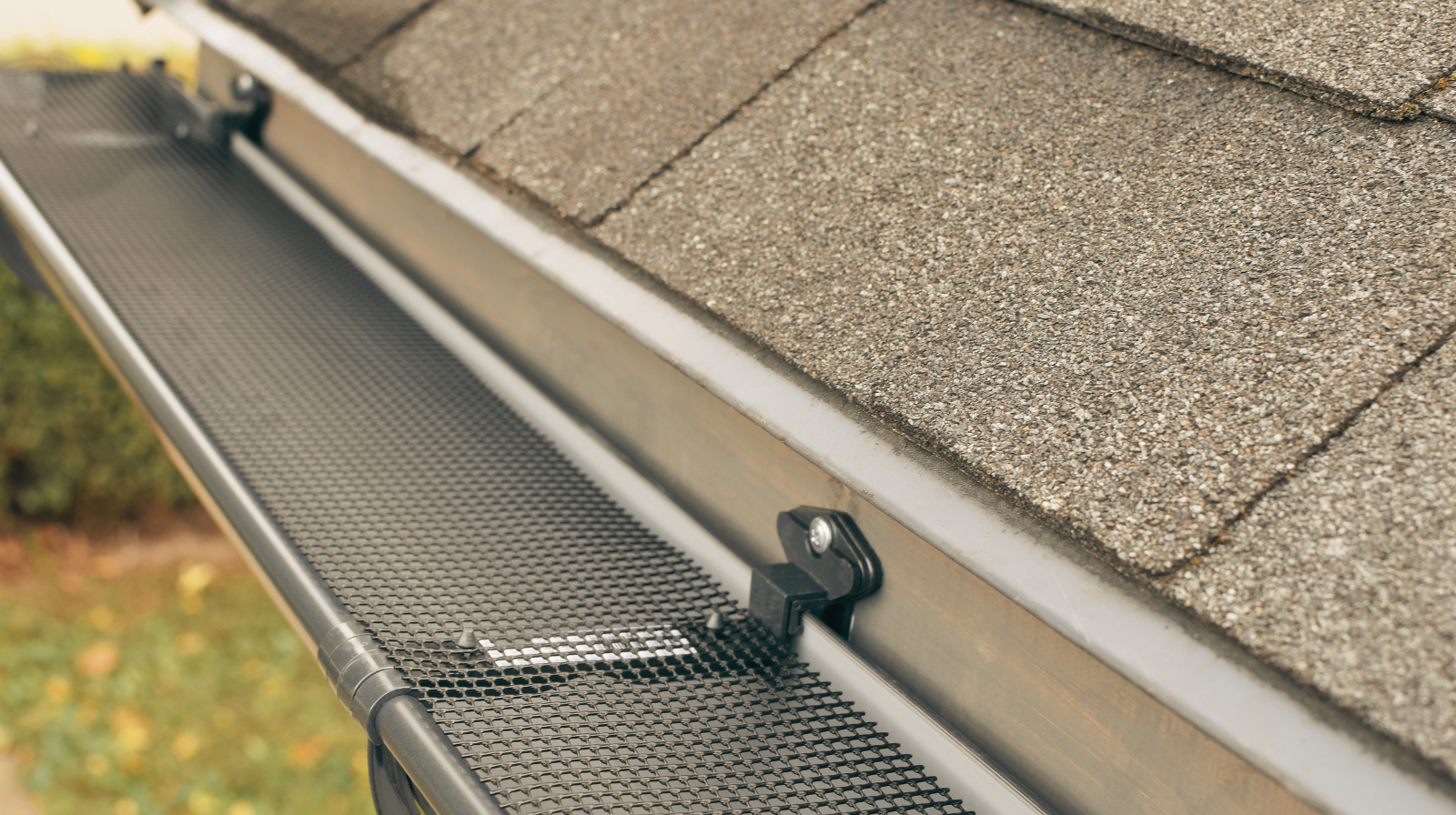 Close-up of a house rain gutter with a black mesh gutter guard installed under asphalt roof shingles.