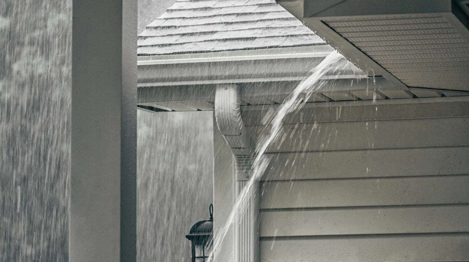Rainwater rushing off a sideways roof gutter, splashing down during heavy rain on a gray house exterior.
