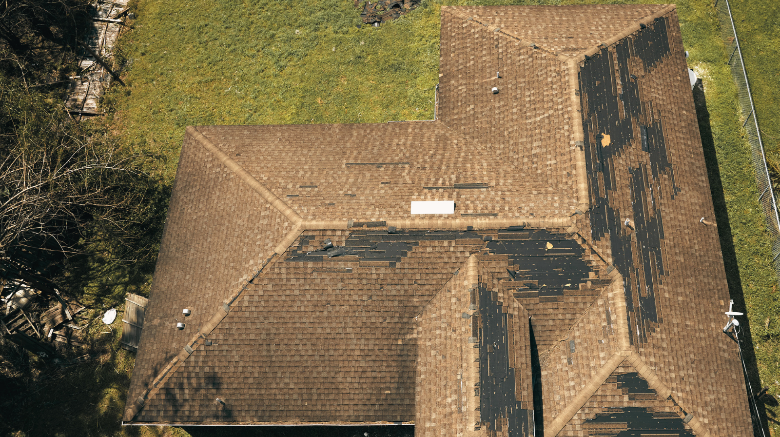 Aerial view of a brown shingle roof with extensive storm damage showing missing shingles and exposed black underlayment.