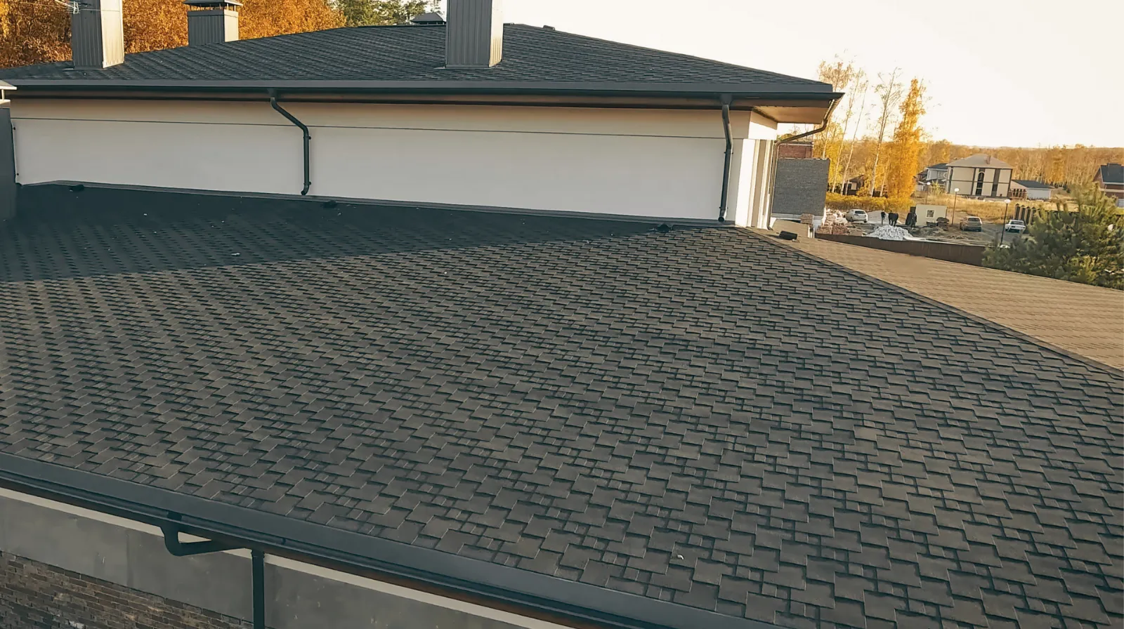 Close-up of a dark shingle roof on a modern house with trees and other houses in the background during autumn.