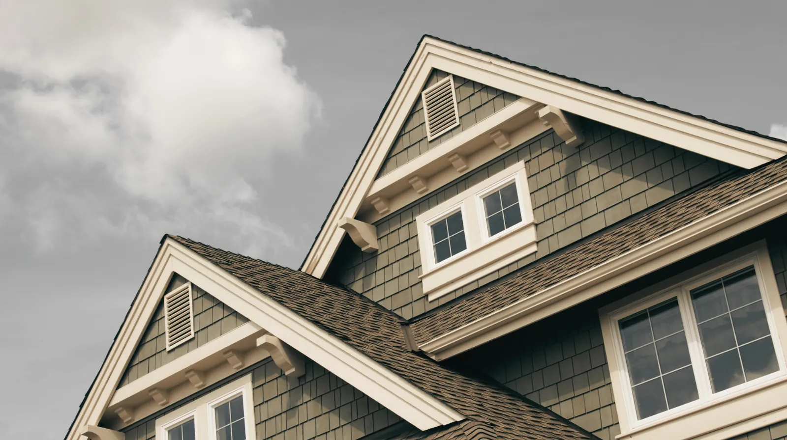 Close-up of a house roof with brown shingles and gray siding under a partly cloudy sky.