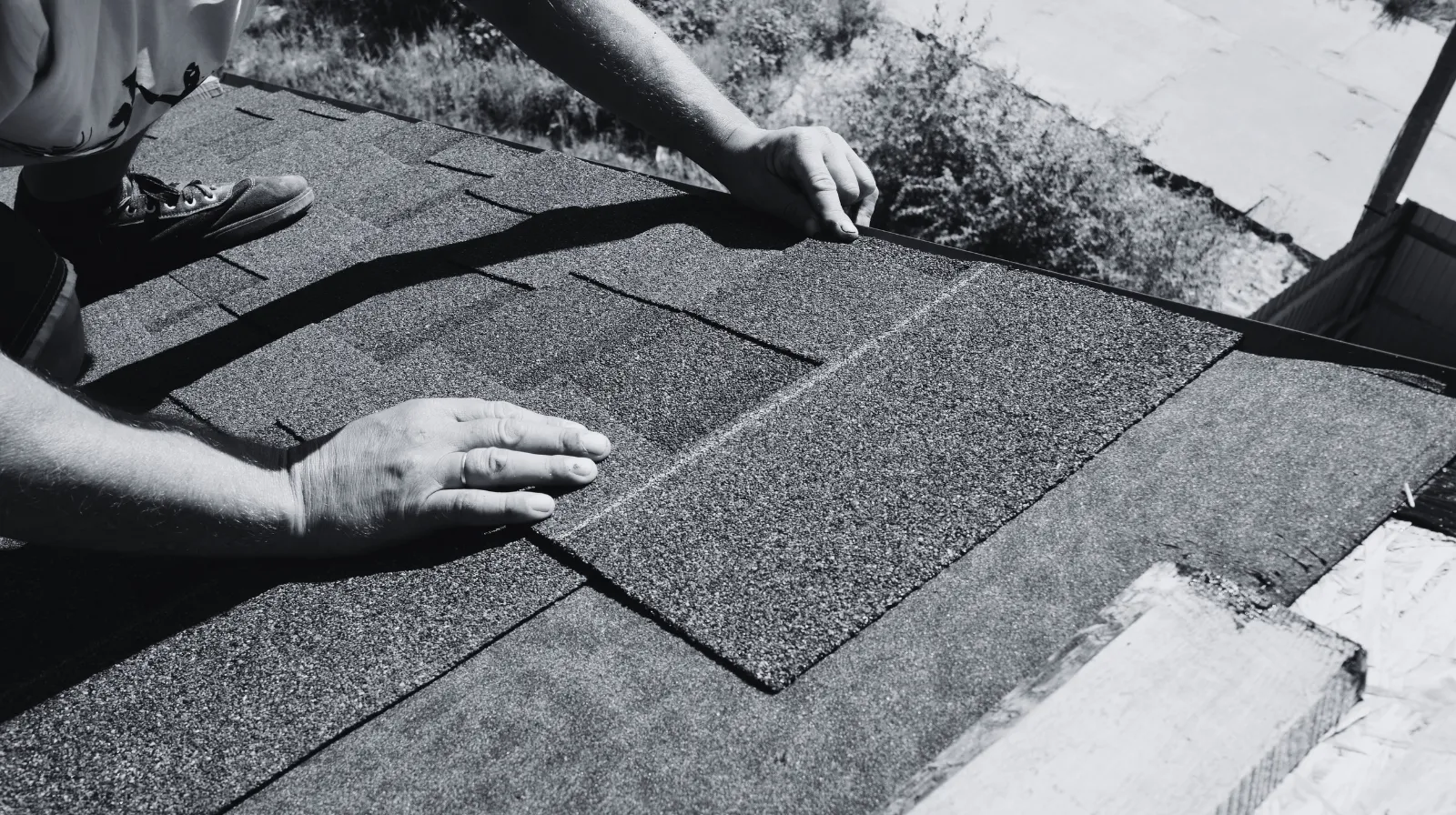 Person installing asphalt roofing shingles on a roof in black and white.