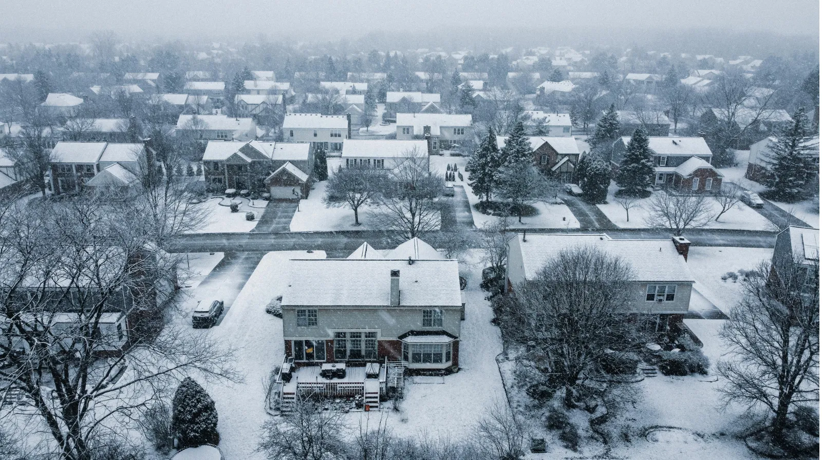 Snow-covered suburban neighborhood with houses, trees, and roads during a snowfall.