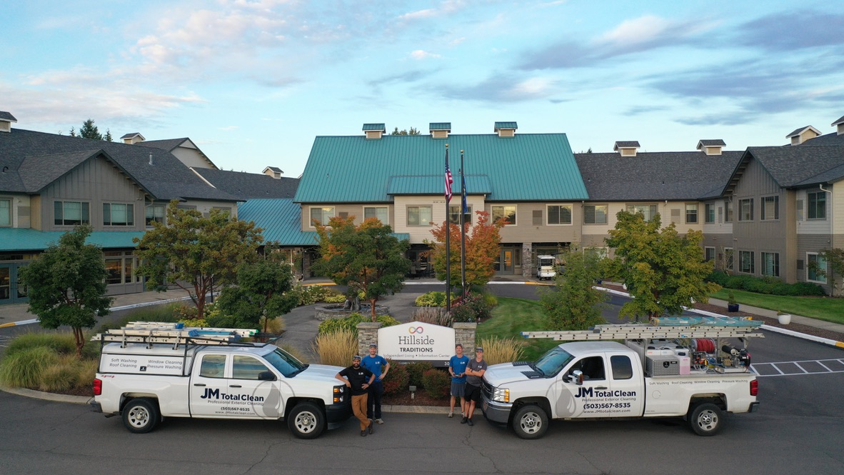 Two white JM Total Clean trucks parked in front of Hillside Traditions building with four workers standing beside the trucks and American flag in the center.