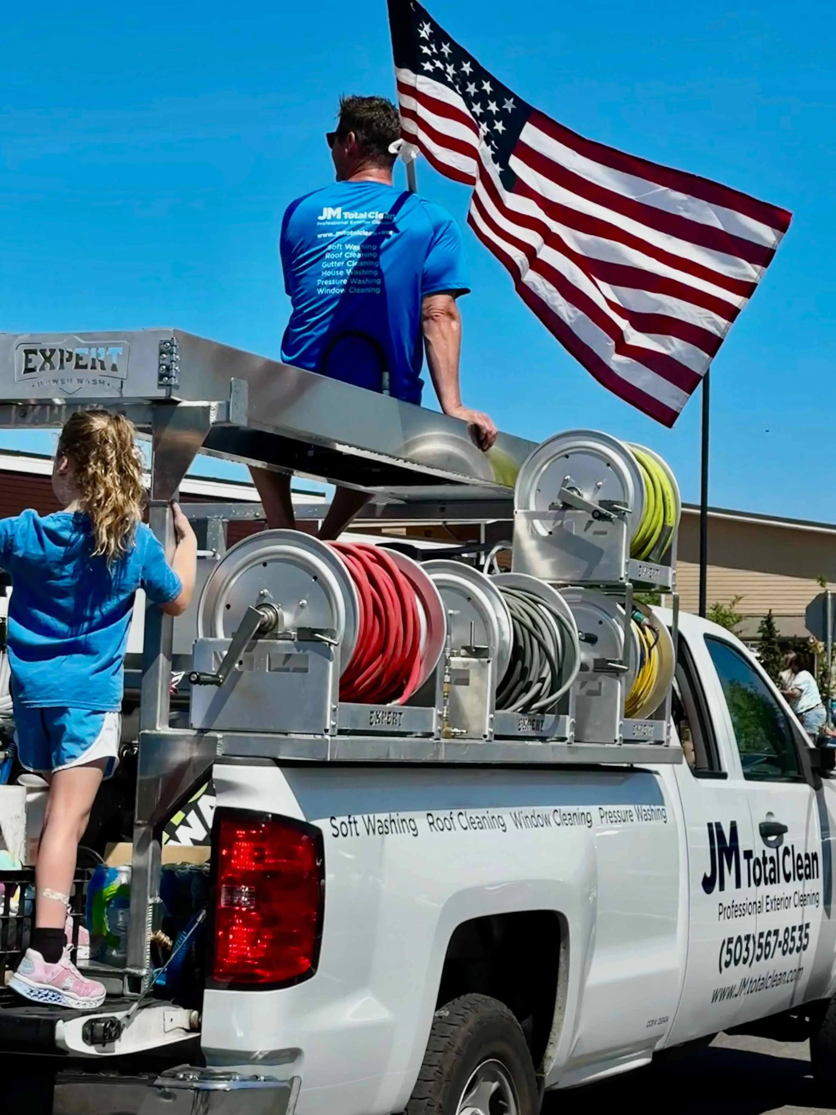 Man and girl on white JM Total Clean truck with hose reels and American flag waving.