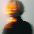 Blurred portrait of a person with short hair and orange light streaks across their face against a gray background.