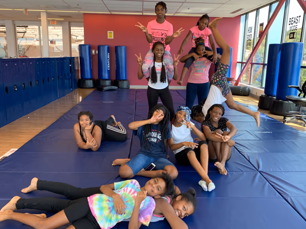 Group of smiling girls in colorful clothes posing on blue gym mats in a room with large windows and lockers.