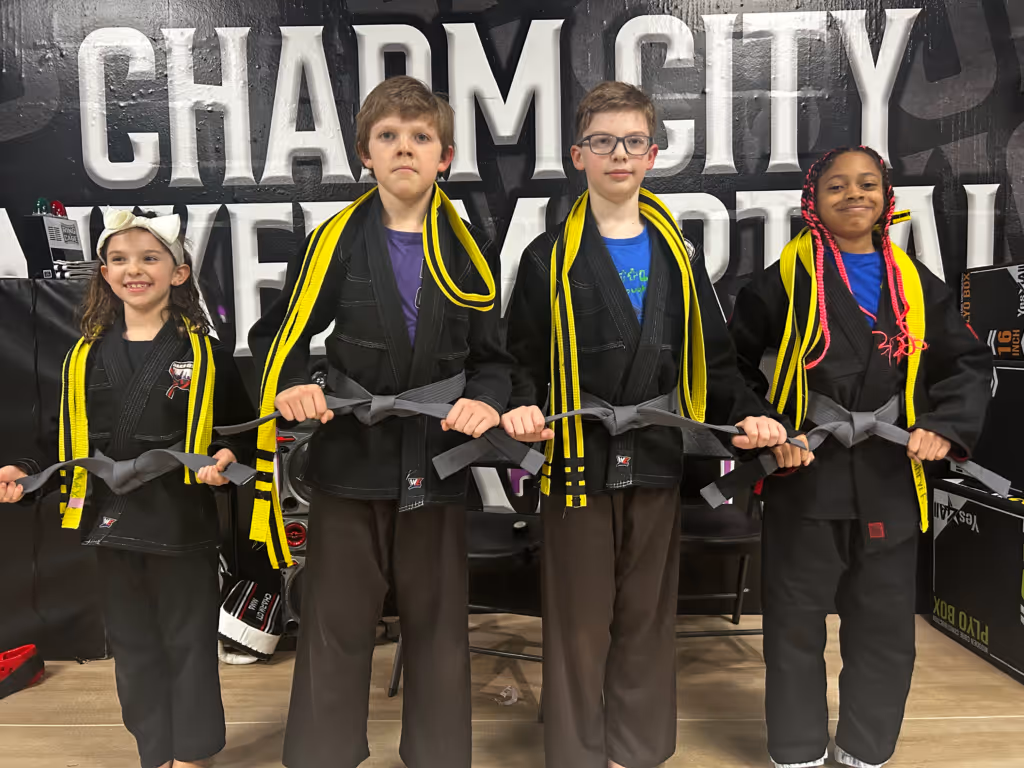Four children in black martial arts uniforms holding gray belts with yellow-striped belts over their shoulders, standing in front of a Charm City Brazilian Jiu-Jitsu banner.