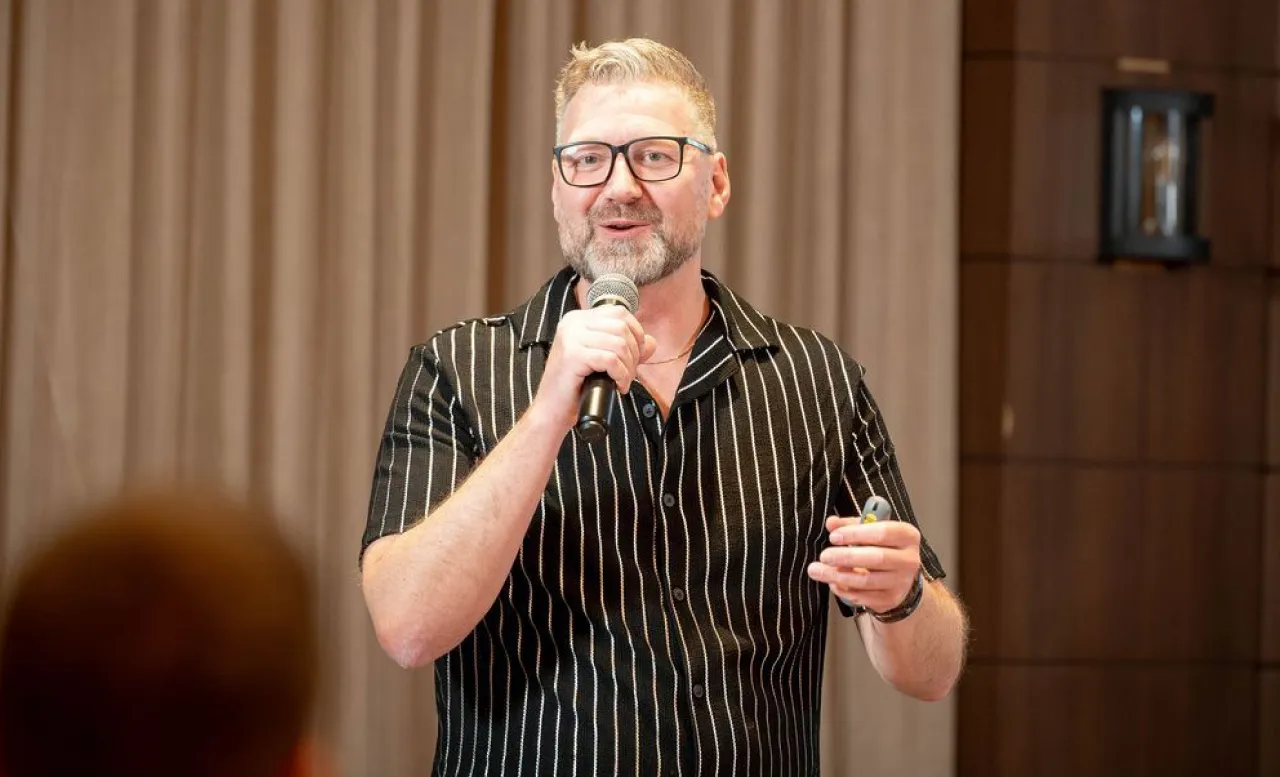 Middle-aged man with glasses and a beard speaking into a microphone during a presentation, wearing a black shirt with white vertical stripes.