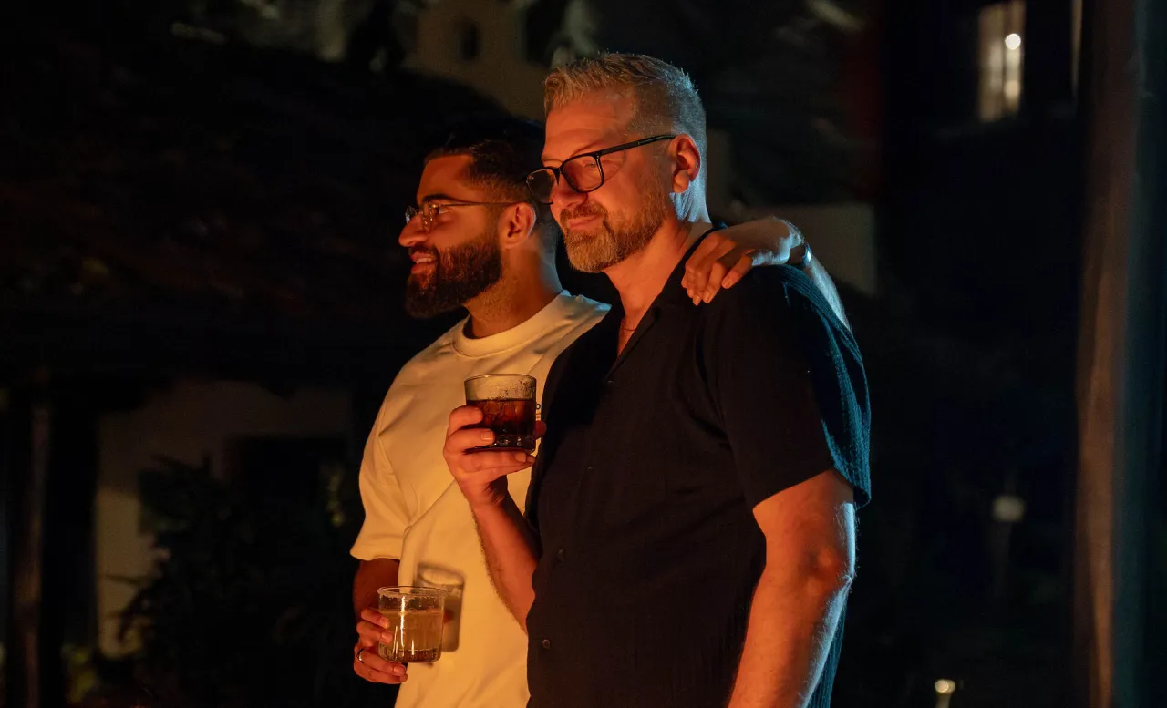 Two men with glasses standing close together and smiling, each holding a glass of drink, in a dimly lit outdoor setting at night.
