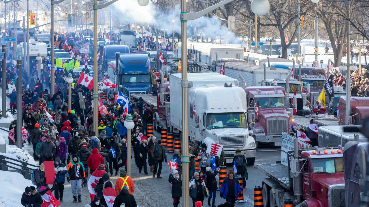 Crowd of people and large trucks with Canadian flags gathered on a snowy city street during a protest or rally.