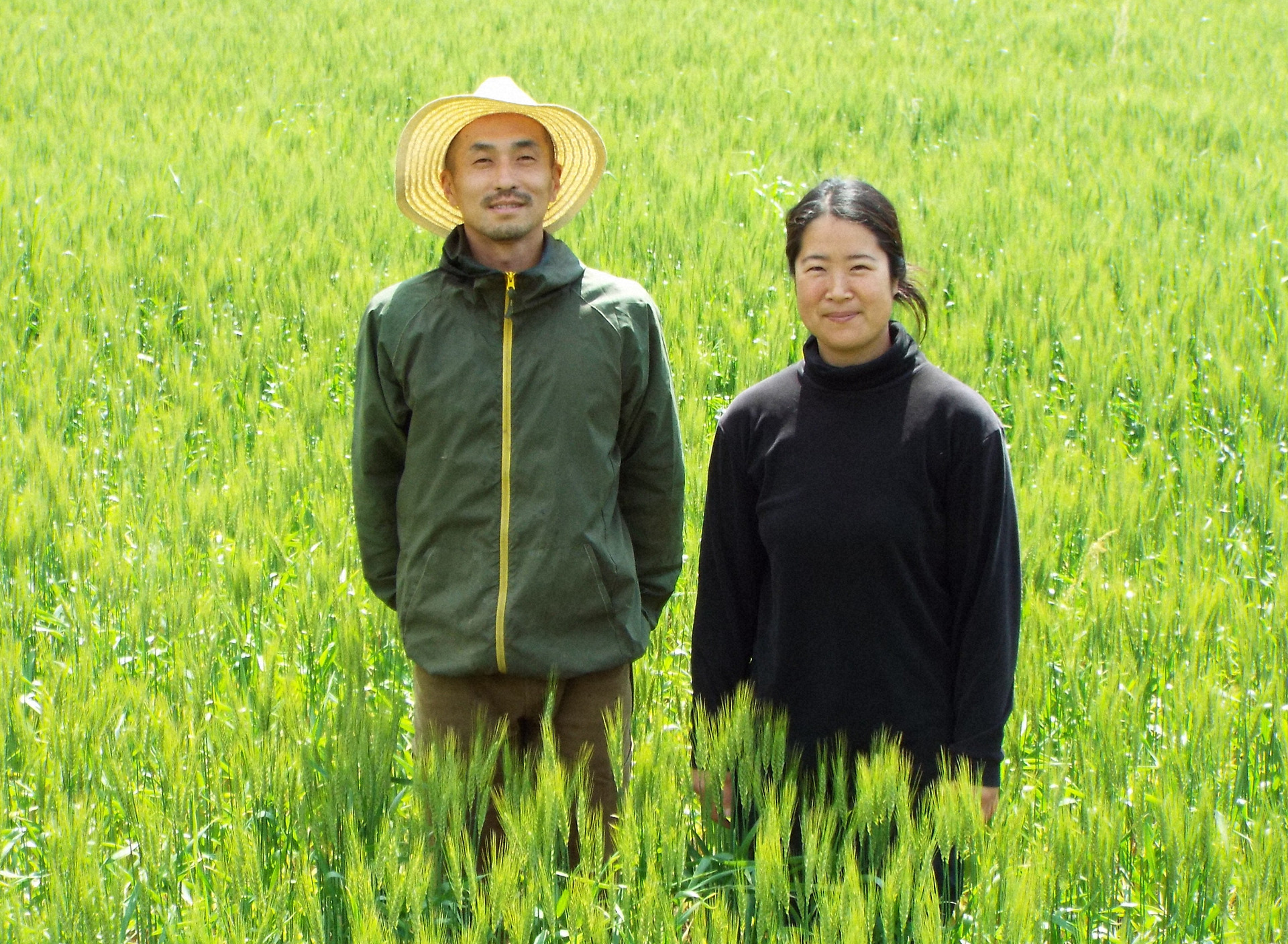 麦の圃場に立つ吉田さん夫妻。「今、青葉が一番美しい季節」とほほ笑む
